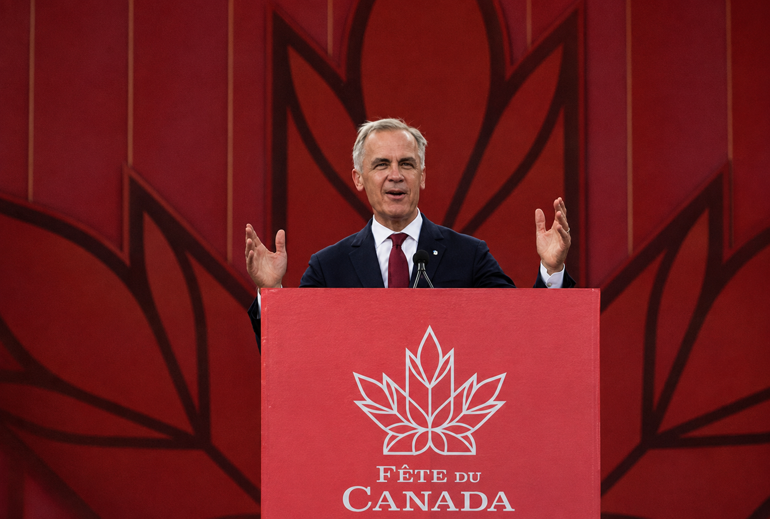 Allocution officielle au pupitre de la Fête du Canada, devant un décor rouge orné de feuilles d’érable.
